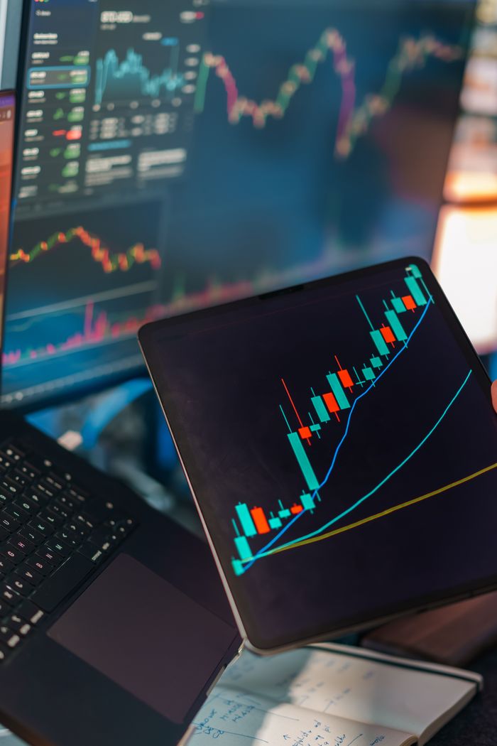 about-us A tablet displaying a candlestick chart with a laptop screen showing stock market data for financial analysis.